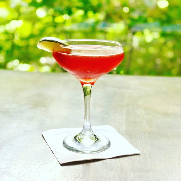 Red cocktail in a coupe glass with a lime wedge, on a white napkin and wooden table.