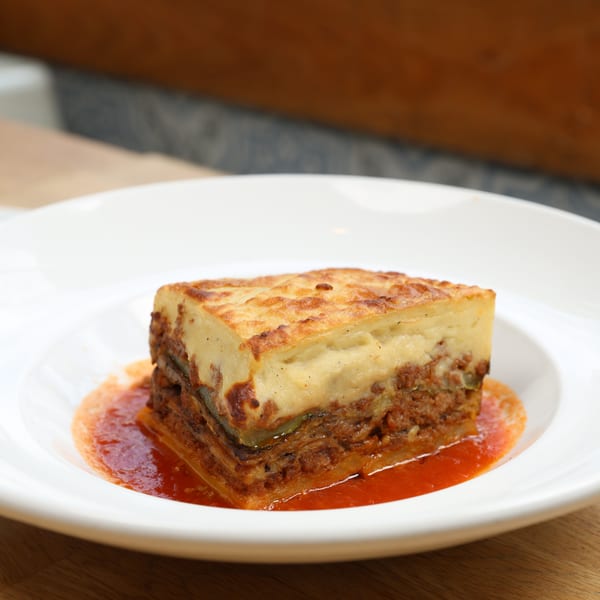 A square slice of moussaka with a golden-brown top, served in a shallow white bowl with red sauce.