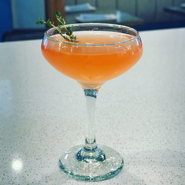A vibrant orange cocktail in a coupe glass, garnished with a sprig of thyme on a white speckled surface.
