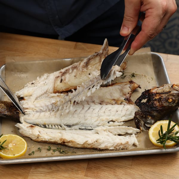 Hands using tongs to debone a whole grilled fish on a baking sheet with lemon and rosemary.