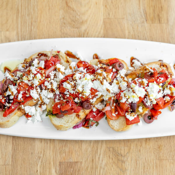 Four bruschetta pieces topped with diced tomatoes, feta, olives, and balsamic glaze on a white plate.