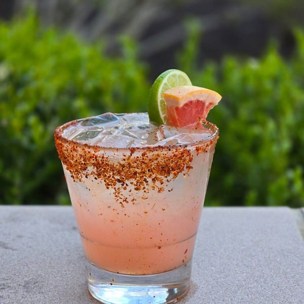 Pink cocktail with ice, a chili-lime rim, and garnished with lime and grapefruit slices.