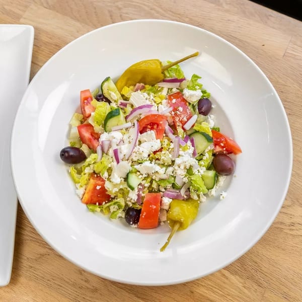 A vibrant Greek salad with lettuce, tomatoes, cucumbers, feta, olives, and pepperoncini on a white plate.