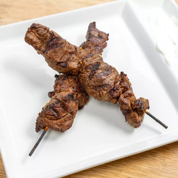Two grilled beef skewers with char marks on a white rectangular plate with a side of tzatziki.