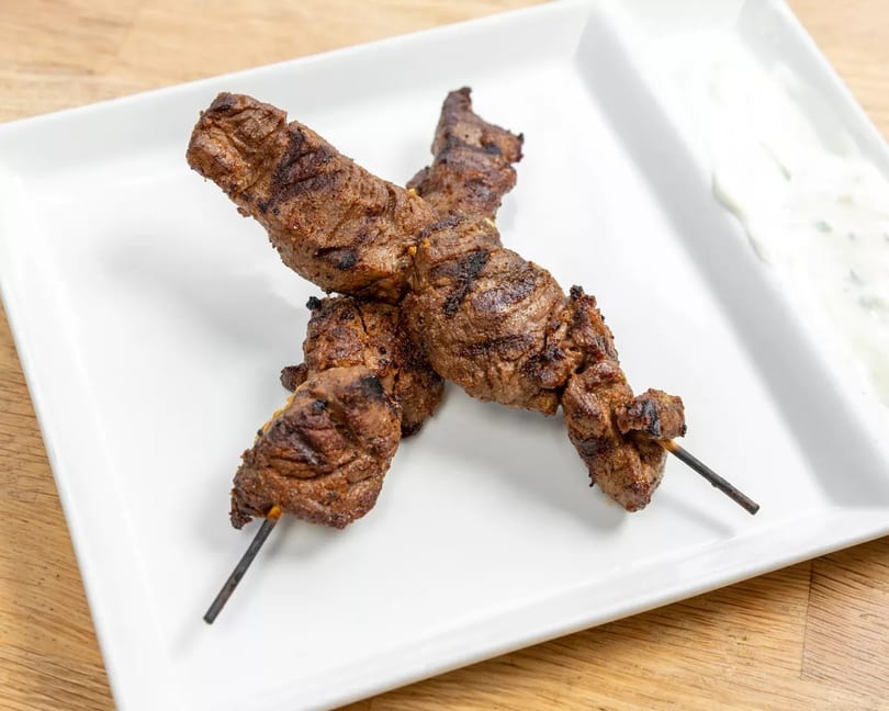 Two grilled beef skewers with char marks on a white rectangular plate with a side of tzatziki.