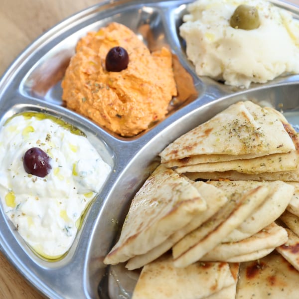 A metal platter with four compartments holding pita bread, tzatziki, hummus, and mashed potatoes.
