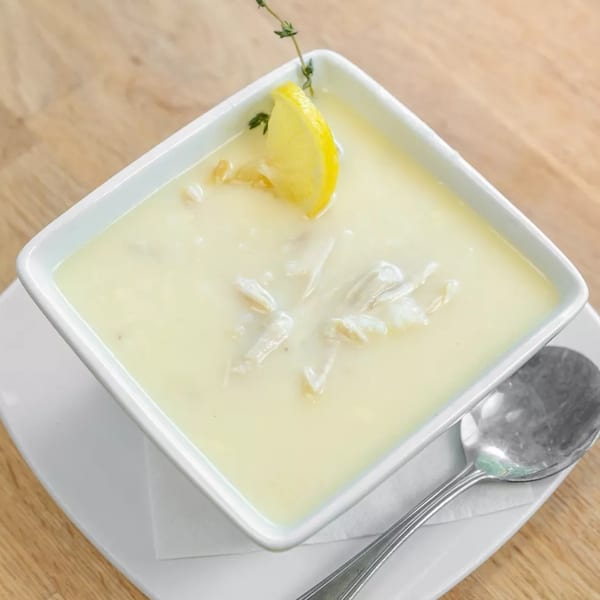 Creamy avgolemono soup with chicken, garnished with a lemon slice and thyme, on a wooden table.