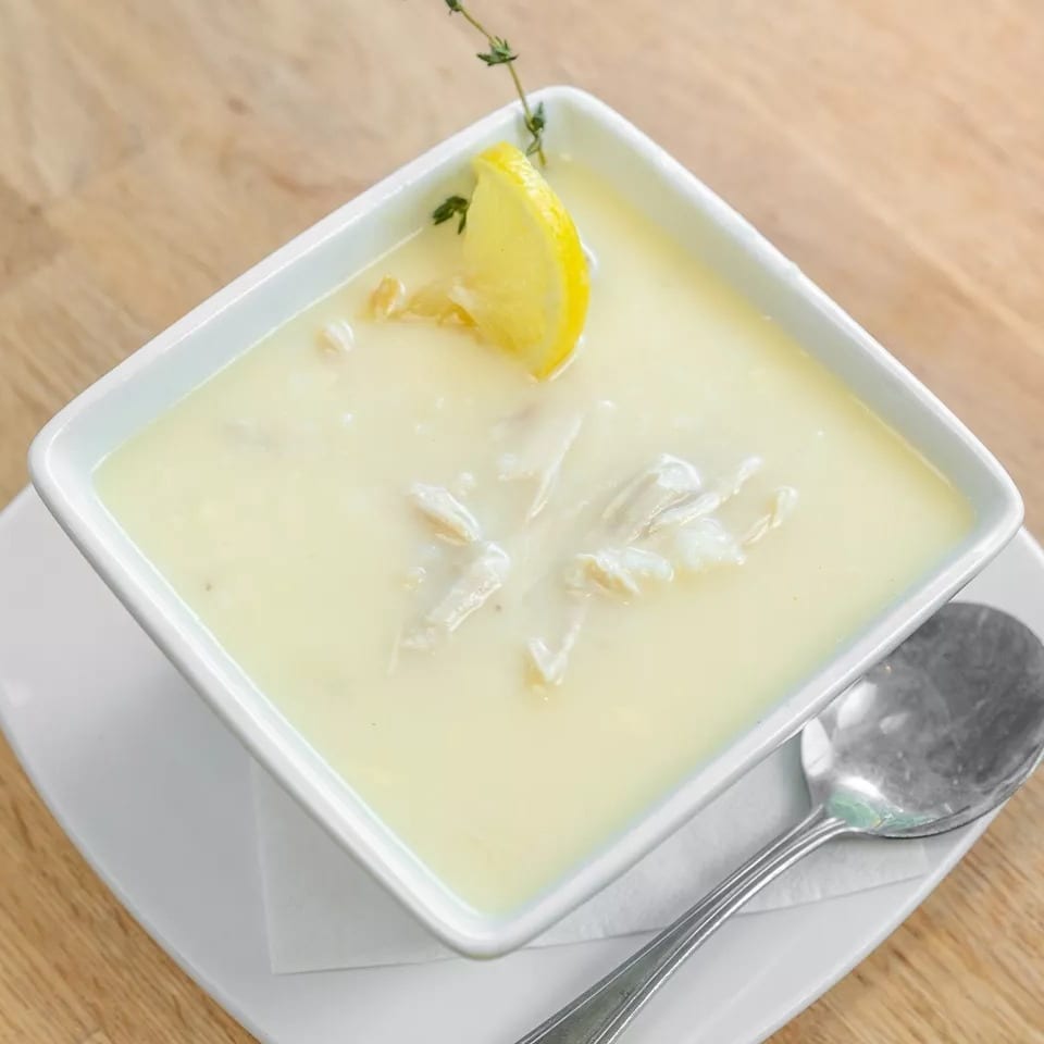 Creamy avgolemono soup with chicken, garnished with a lemon slice and thyme, on a wooden table.